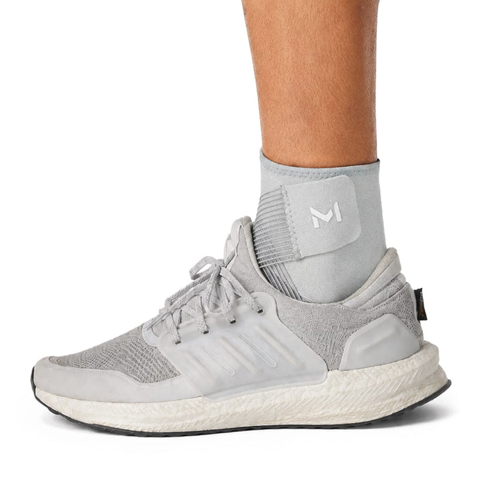 En person har en grå sneaker på og en lysegrå justerbar ankelstøtte med et hvidt M-logo.