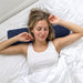 En kvinde i en hvid tanktop sidder afslappet og smiler, mens hun hviler hovedet på en ergonomisk nakkepude i memoryfoam.