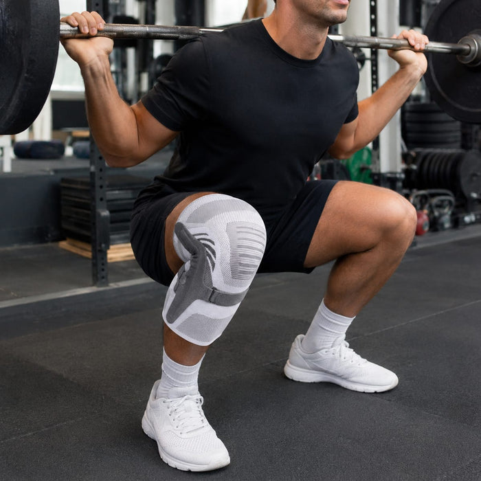 En mand i sort tøj udfører squats med vægtstang i fitnesscenteret, iført en knæstøtte med spænde fra Kroppsbutiken.